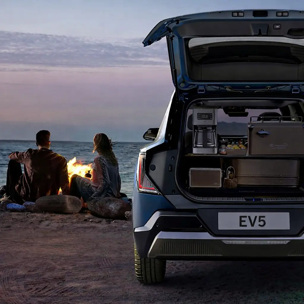 A Kia EV5 electric SUV with tail gate opened in a beach with a couple and bon fire